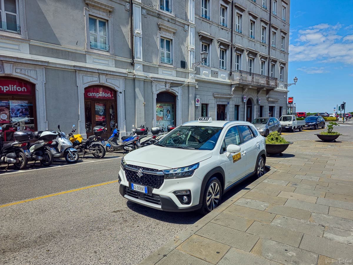 White taxi parked on a city street beside scooters and a historic stone building, representing typical transport options. A common and convenient choice for getting a taxi from a Greek airport to your hotel, especially if you're carrying luggage or arriving late.