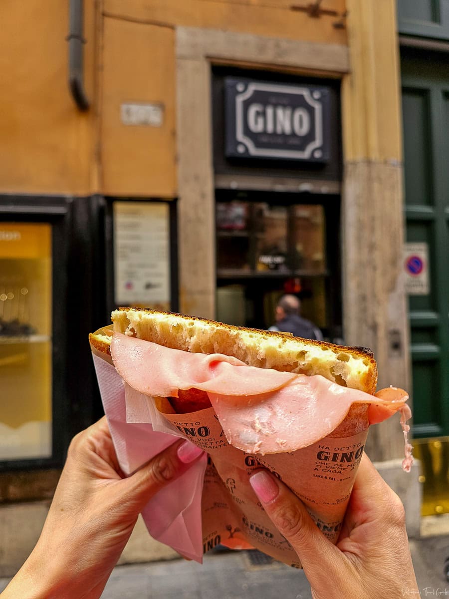 A close-up of a pizza bianca con mortadella held in front of Da Gino, a popular Roman street food spot. The fluffy, golden focaccia-style bread is stuffed with thin slices of silky mortadella, wrapped in branded paper for a classic and simple Roman snack.
