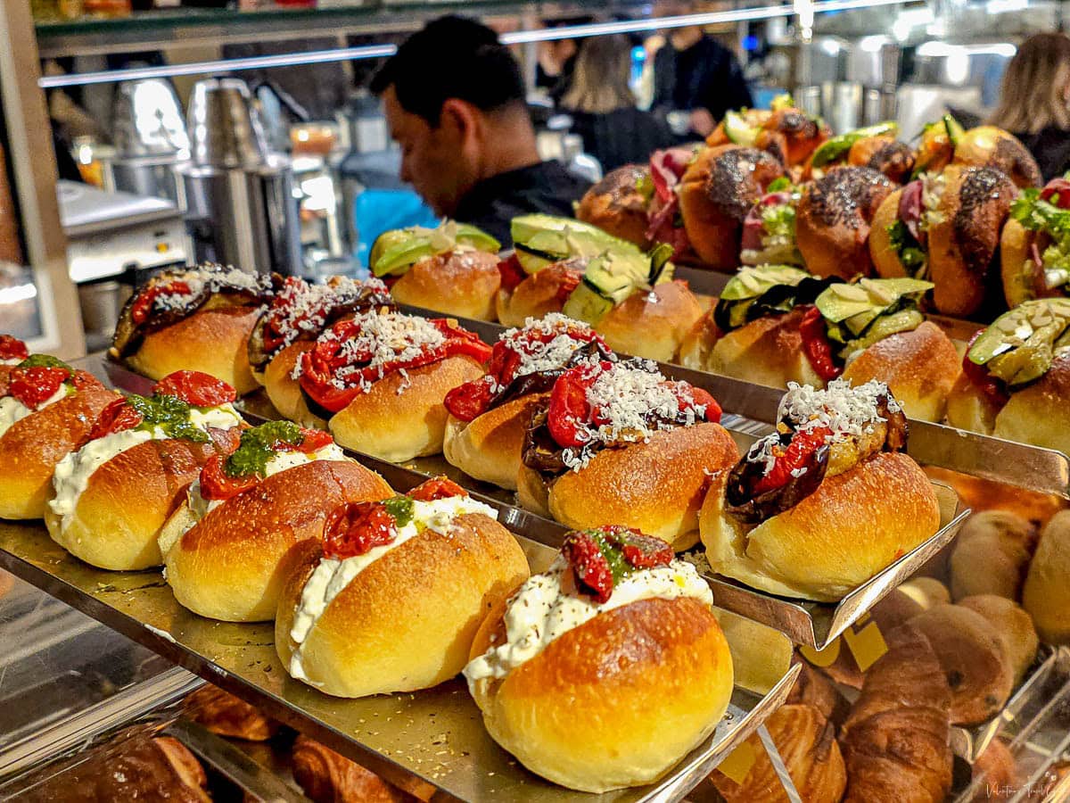 A display of savory maritozzi—soft, golden brioche buns filled with ingredients like whipped ricotta, roasted cherry tomatoes, grilled zucchini, pesto, and grated cheese—arranged in neat rows at a bustling counter. The rich variety of colors and textures highlights a modern twist on the classic Roman pastry, traditionally served sweet.
