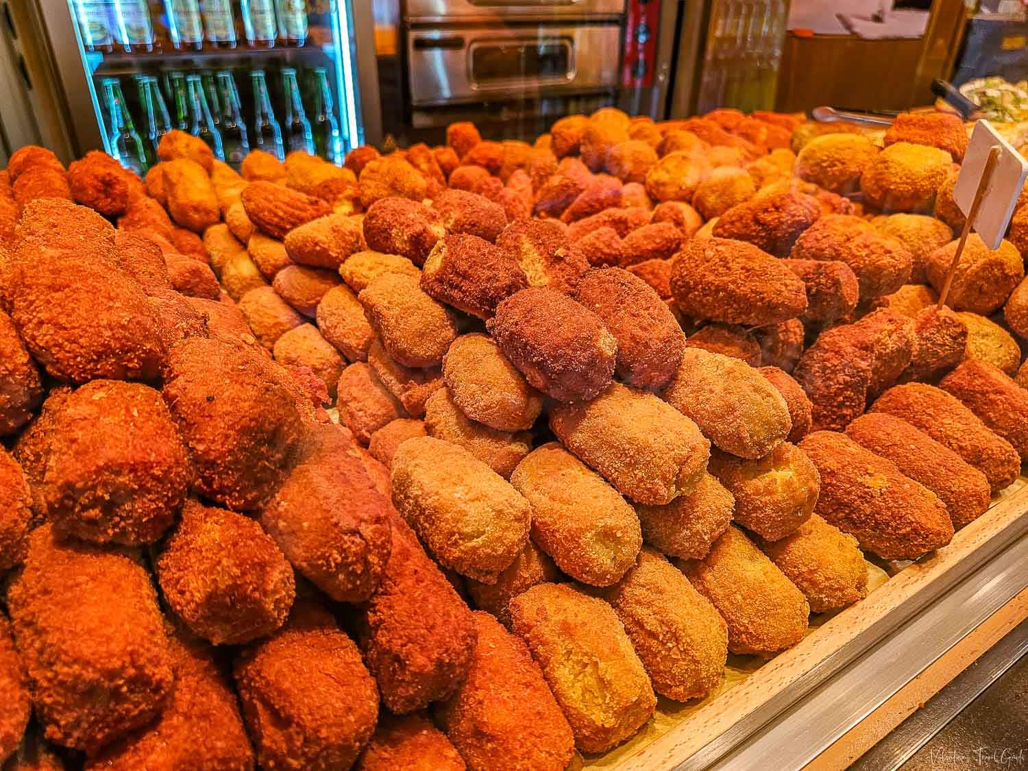 A mouthwatering display of freshly fried supplì, crispy, golden-brown rice croquettes, stacked high behind a glass counter in a Roman food shop. The variety of colors hints at different fillings, with their crunchy coating promising a gooey, savory surprise inside.
