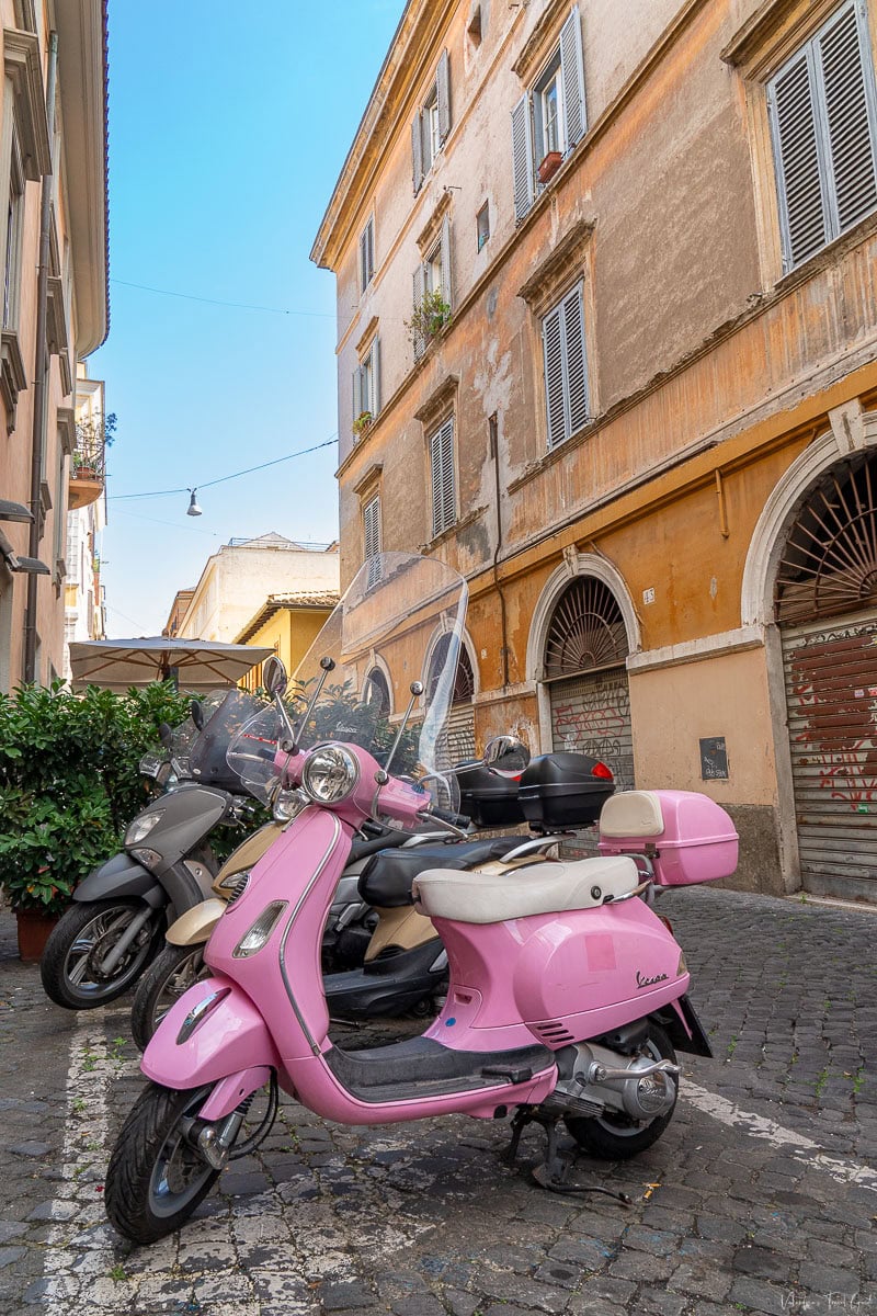 A bright pink Vespa scooter is parked in a narrow cobblestone alley in Rome, surrounded by other scooters and rustic, shuttered buildings. A fun reminder of don’t assume riding a scooter is the easiest option without experience; Rome’s traffic, tight spaces, and aggressive driving can make it risky for unseasoned riders.