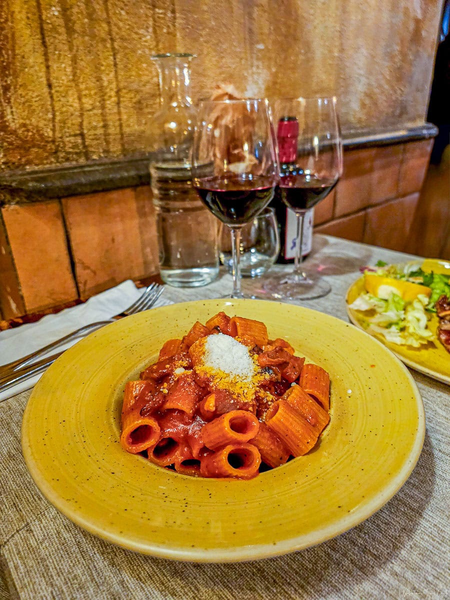 Rigatoni all’Amatriciana served on a rustic yellow plate, paired with red wine at a cozy trattoria—perfect for those looking to book a restaurant for authentic Italian cuisine