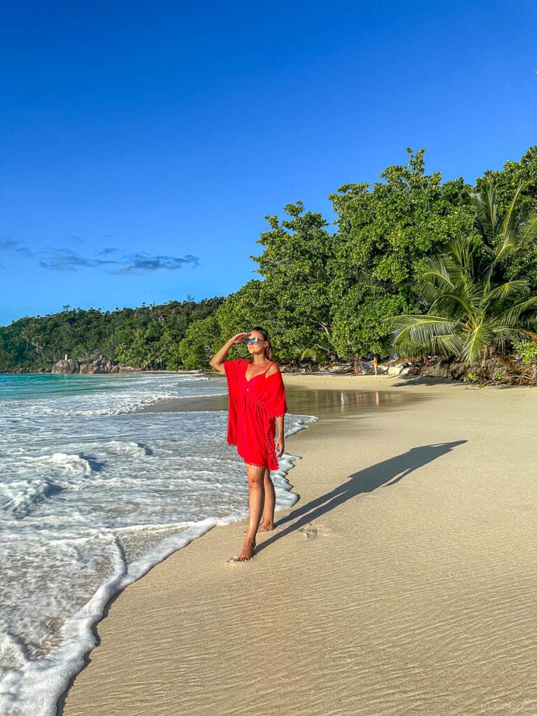 Valentina strolls along the golden shore of Anse Lazio in Praslin, Seychelles, wearing a flowing red beach cover-up under the bright tropical sun—an inspiring scene to introduce natural remedies for a sunburn after sun-soaked adventures.