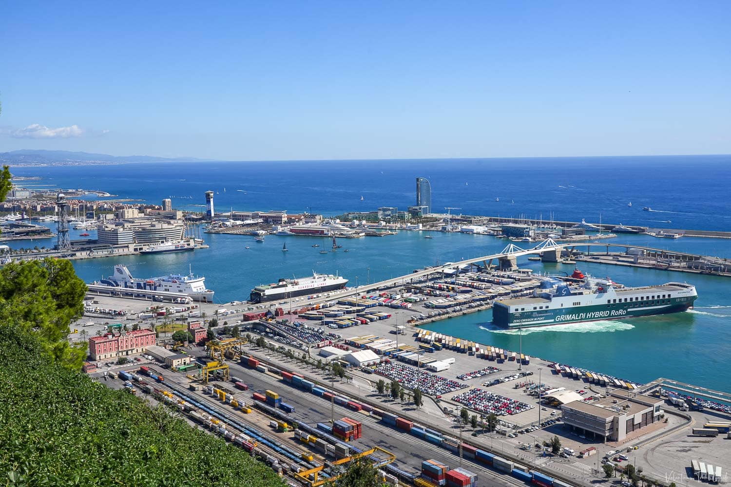 Wide-angle view of the bustling cruise port of Barcelona, featuring several large cruise and ferry ships docked at terminal piers, including the Grimaldi Hybrid RoRo vessel in the foreground. The port is surrounded by shipping containers, cars, and infrastructure, with the deep blue Mediterranean Sea and Barcelona skyline stretching out under a clear sky.