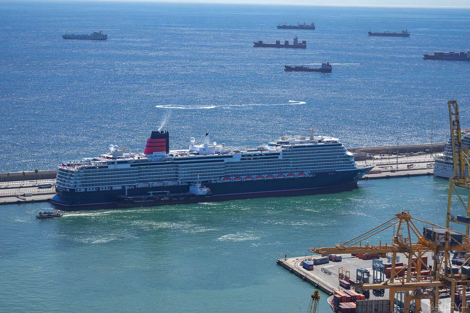 Large cruise ship docked at the terminal in the Port of Barcelona, surrounded by calm blue waters and several cargo ships in the distance, with cranes and containers visible in the foreground. This image captures the busy maritime atmosphere of a cruise Barcelona departure, where modern cruise liners set sail from one of Europe’s most iconic ports.