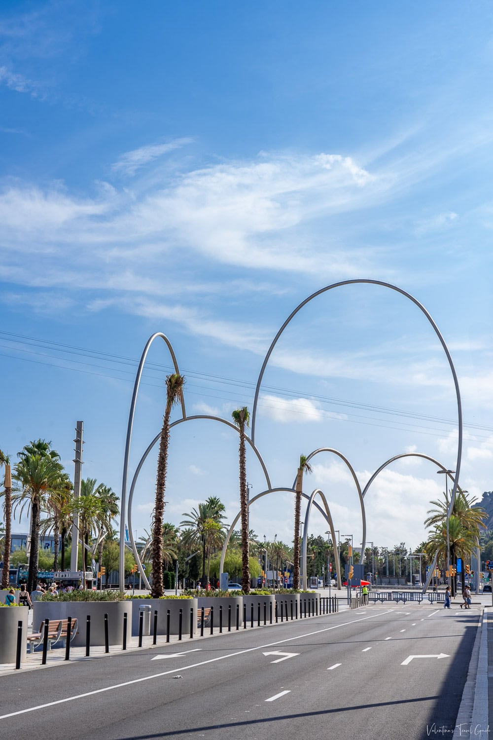 Modern steel arch sculpture at Moll de les Drassanes in Barcelona, surrounded by palm trees, benches, and wide pedestrian walkways on a clear day. This artistic and architectural landmark sits near the Barcelona port cruise area, making it a scenic spot for visitors arriving or departing from their cruise journey.