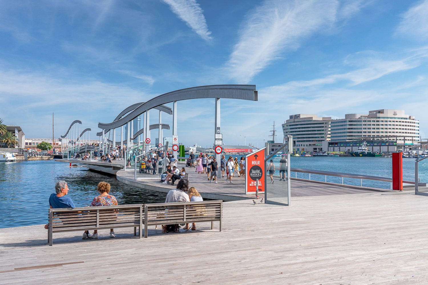 Sunny scene at Rambla de Mar in Barcelona, a pedestrian bridge with flowing architectural curves connecting to Maremagnum shopping center, with the World Trade Center and docked boats in the background. This lively walkway is a scenic gateway to Catalonia port Barcelona, blending maritime views with leisure and local culture.