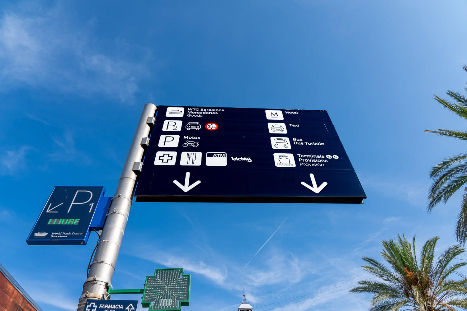 Directional sign near the World Trade Center with icons for parking, taxis, metro, restaurants, and cruise terminals, set against a bright blue sky and palm trees. This wayfinding signage helps guide travelers navigating the Barcelona Spain cruise port, pointing toward key services and cruise terminal access.
